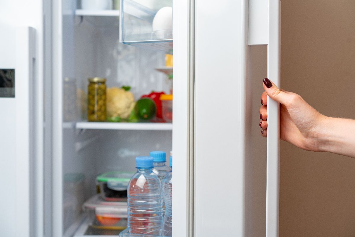 Hand opening a refrigerator stocked with bottled water and fresh food.