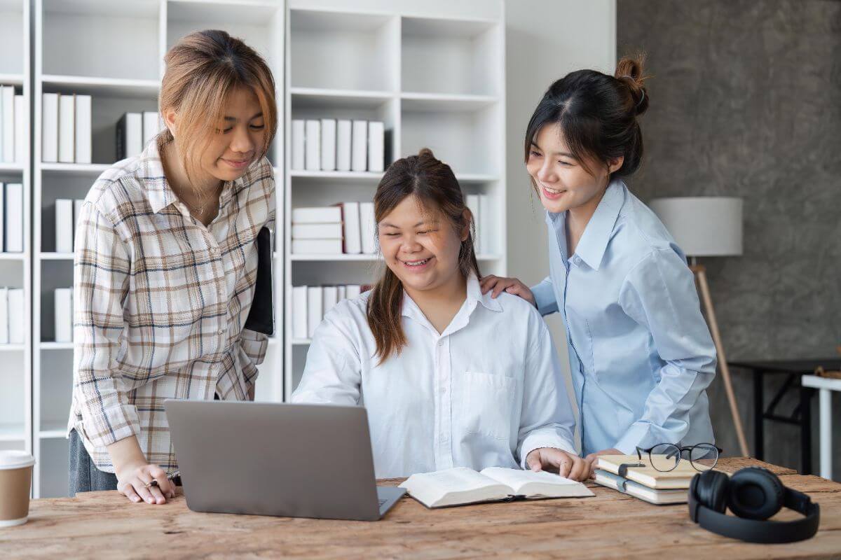 Three women collaborate around a laptop and notebook in an office, showing teamwork and virtual assistant training support.