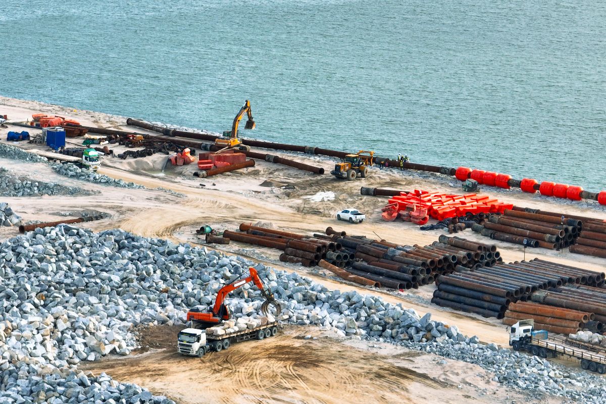 Coastal infrastructure site with steel pipes, heavy equipment, and rock materials near the water