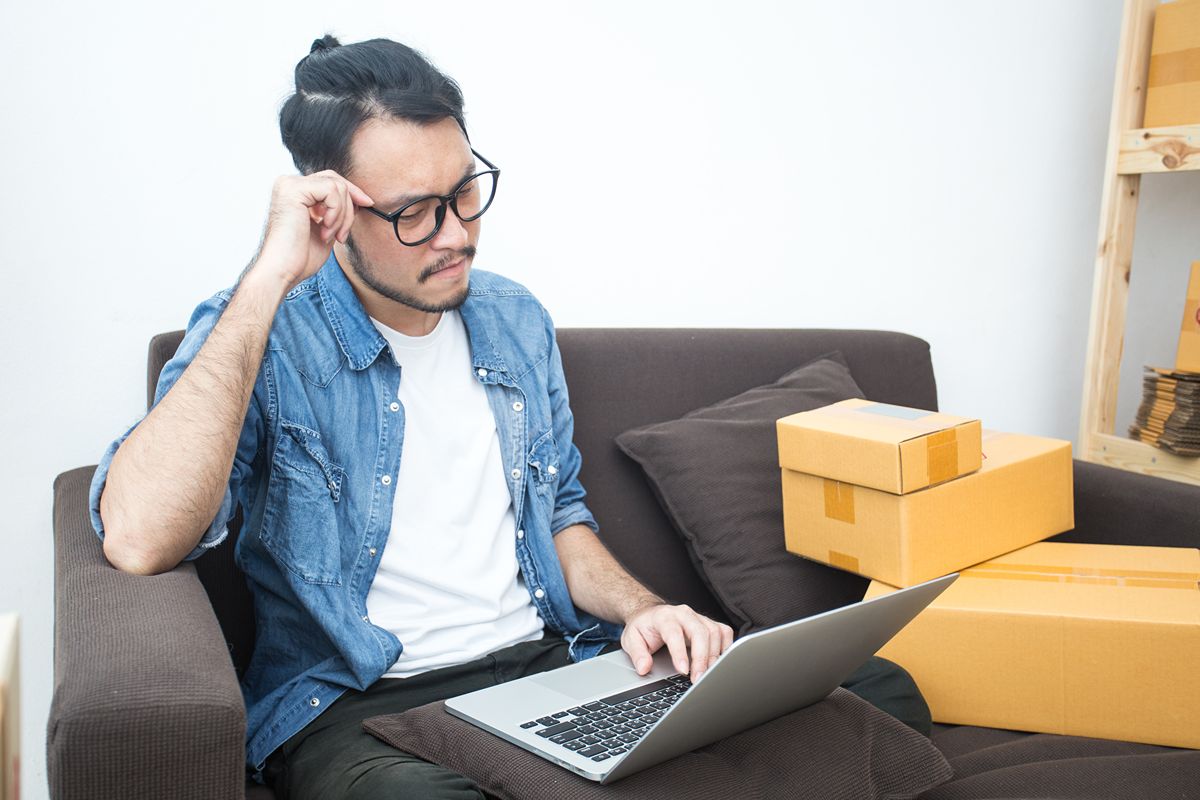 Man shopping for home appliances online using a laptop while seated beside delivered cardboard boxes