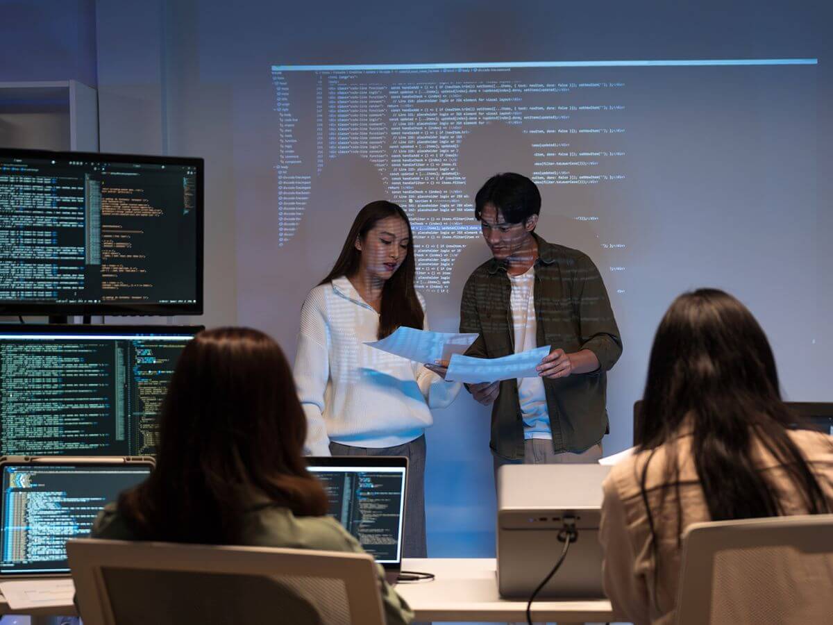 Two students present a programming project to classmates in a computer laboratory with coding displayed on screens